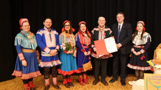 Bildet viser en gruppe på sju personer som står på en scene med svart bakgrunn og tregulv.