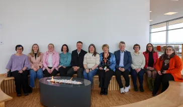 En gruppe på tolv voksne personer sitter på en buet trebenk rundt et rundt ildsted innendørs, i et lyst rom med store vinduer.