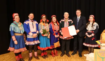 Bildet viser en gruppe på sju personer som står på en scene med svart bakgrunn og tregulv.